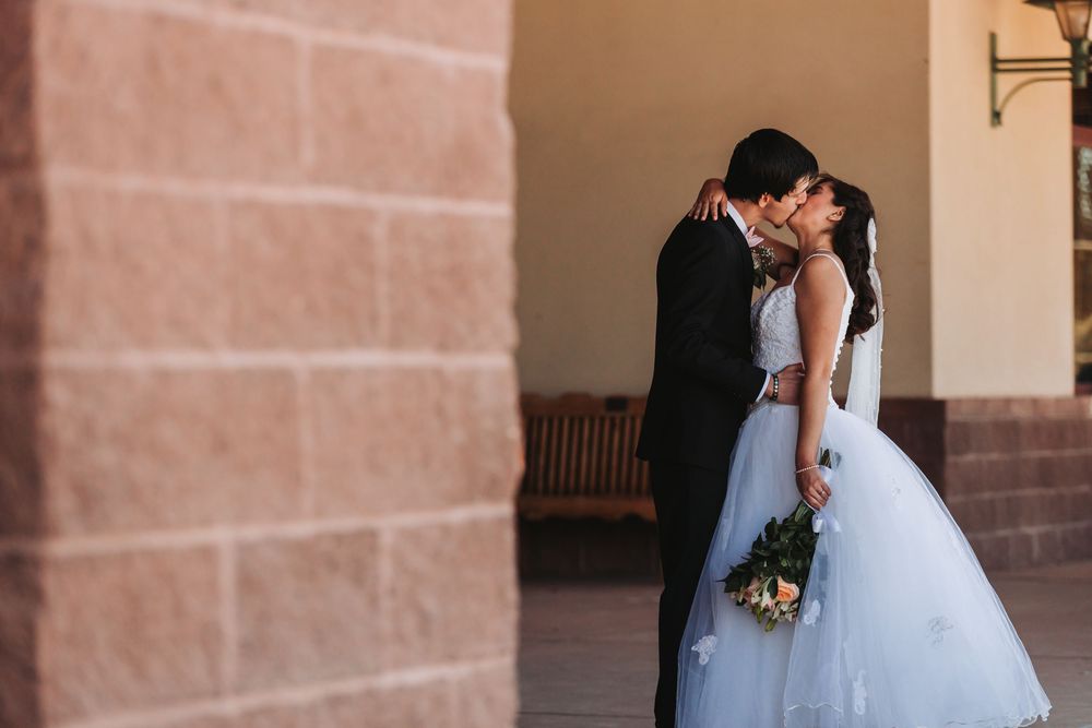 A romantic couple in formal wedding attire embraces and shares a kiss in front of an elegant stone archway.