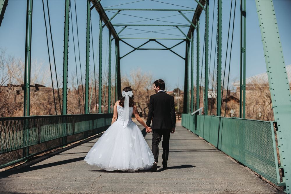 A newlywed couple walks hand in hand across a vintage green metal bridge on their wedding day.