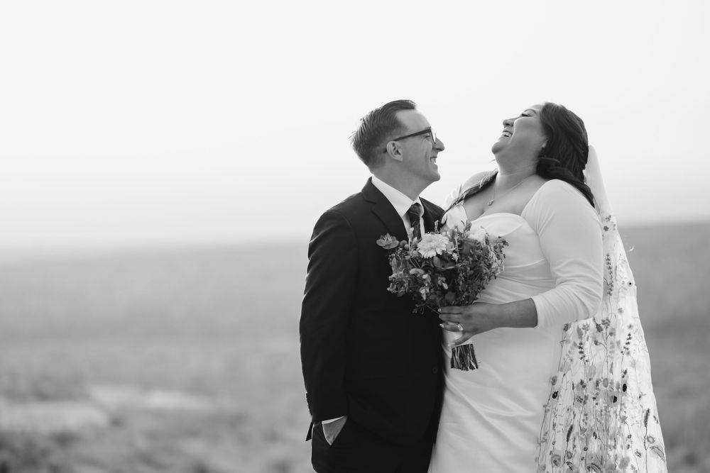 A joyful black and white wedding photo of a couple laughing together while holding a floral bouquet outdoors.