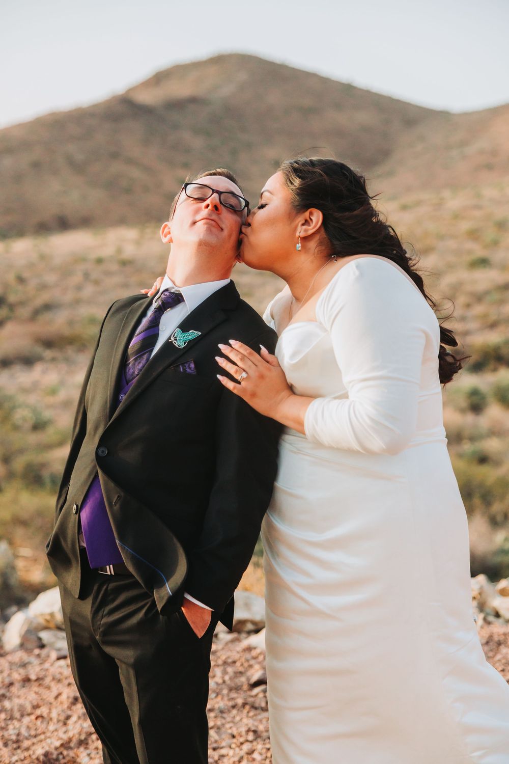 A couple dressed in wedding attire share an intimate moment against a desert mountain backdrop.