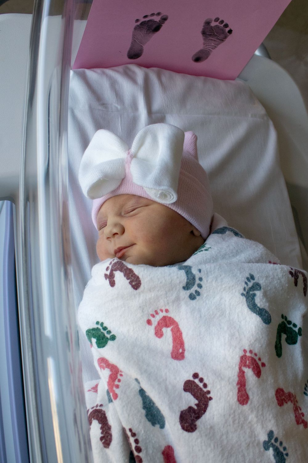Sleeping infant swaddled in white blanket with colorful footprint patterns and wearing light bow hat.