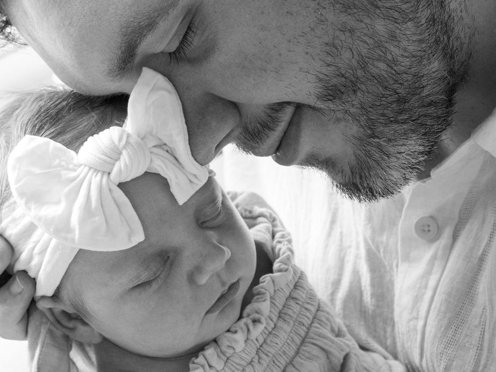 A tender black and white portrait of a new father gently snugging his sleeping baby wearing a white bow headband in Charleston SC.