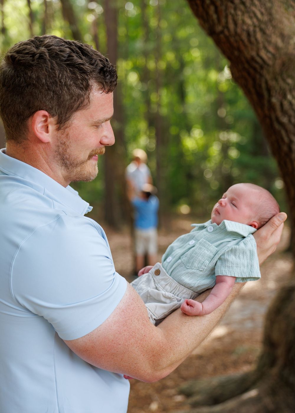 Father holding newborn baby in a sunlit forest, sharing a tender moment outdoors during a family photo session.