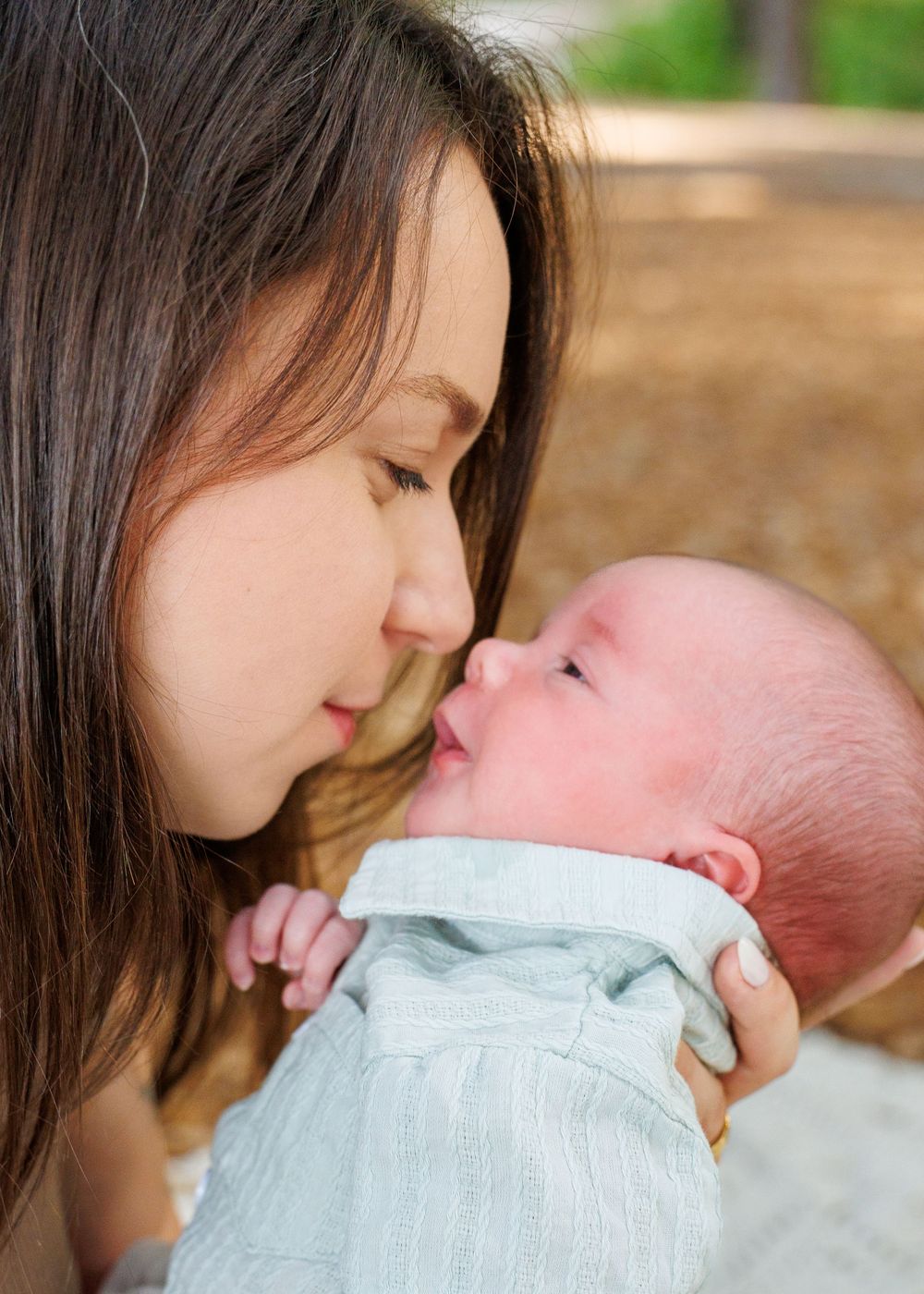 Young mother tenderly holds newborn baby nose-to-nose outdoors in soft natural light.