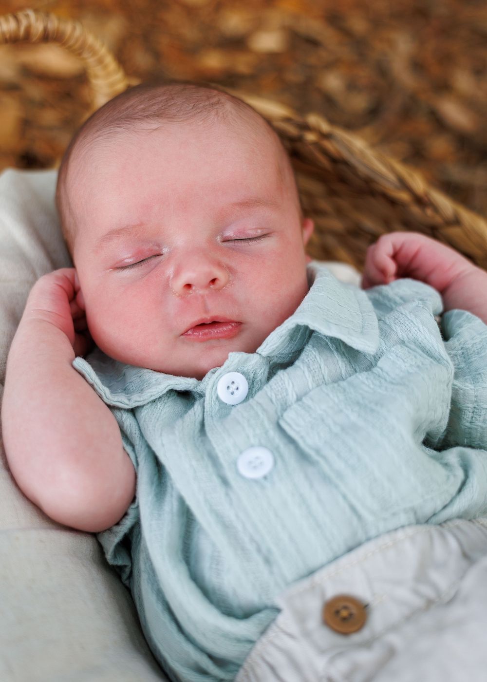 Peaceful newborn baby sleeping in a wicker basket wearing a soft mint green romper with white buttons.