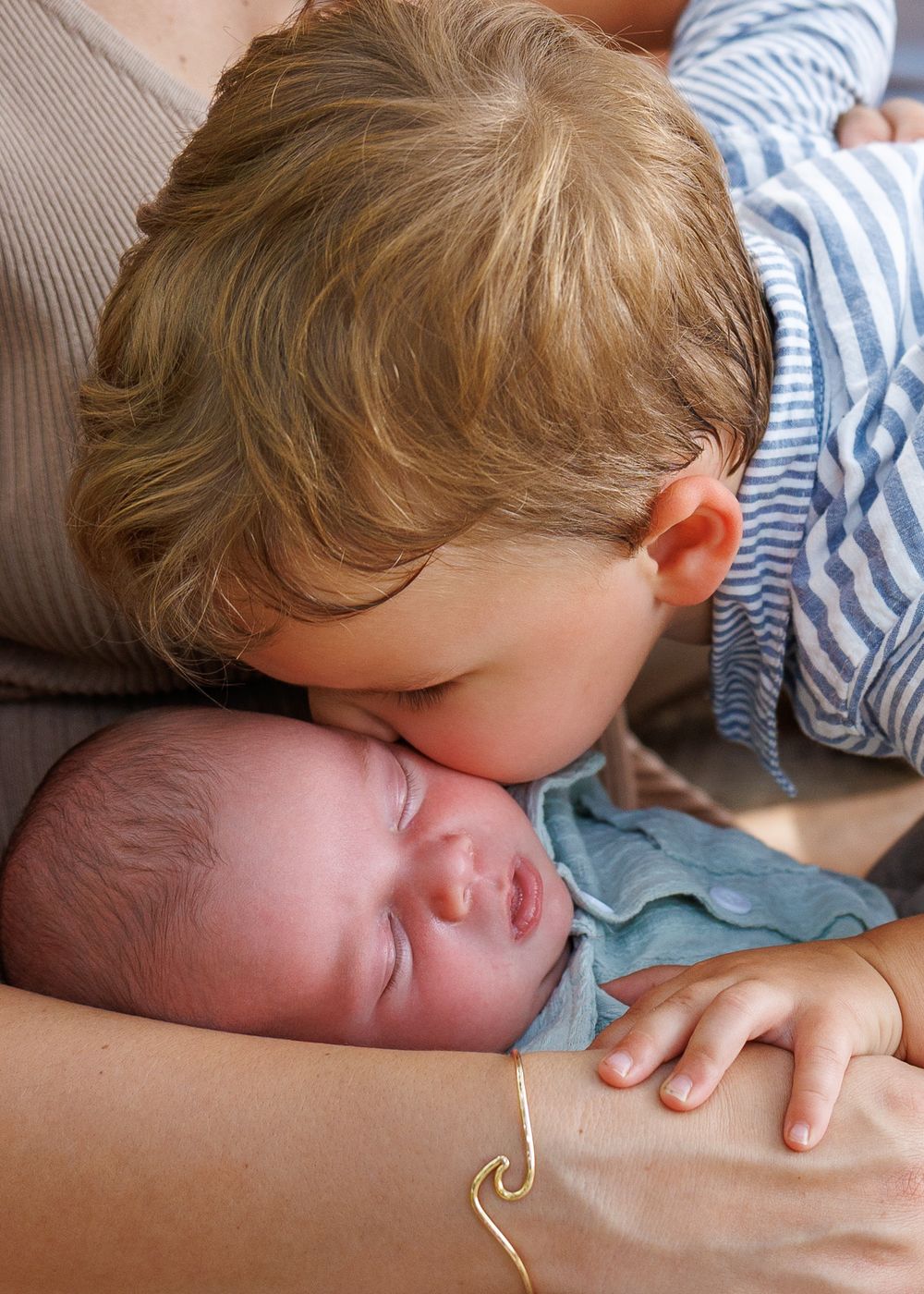 Young boy gently kissing sleeping newborn baby sibling, tender family moment captured in warm natural light.