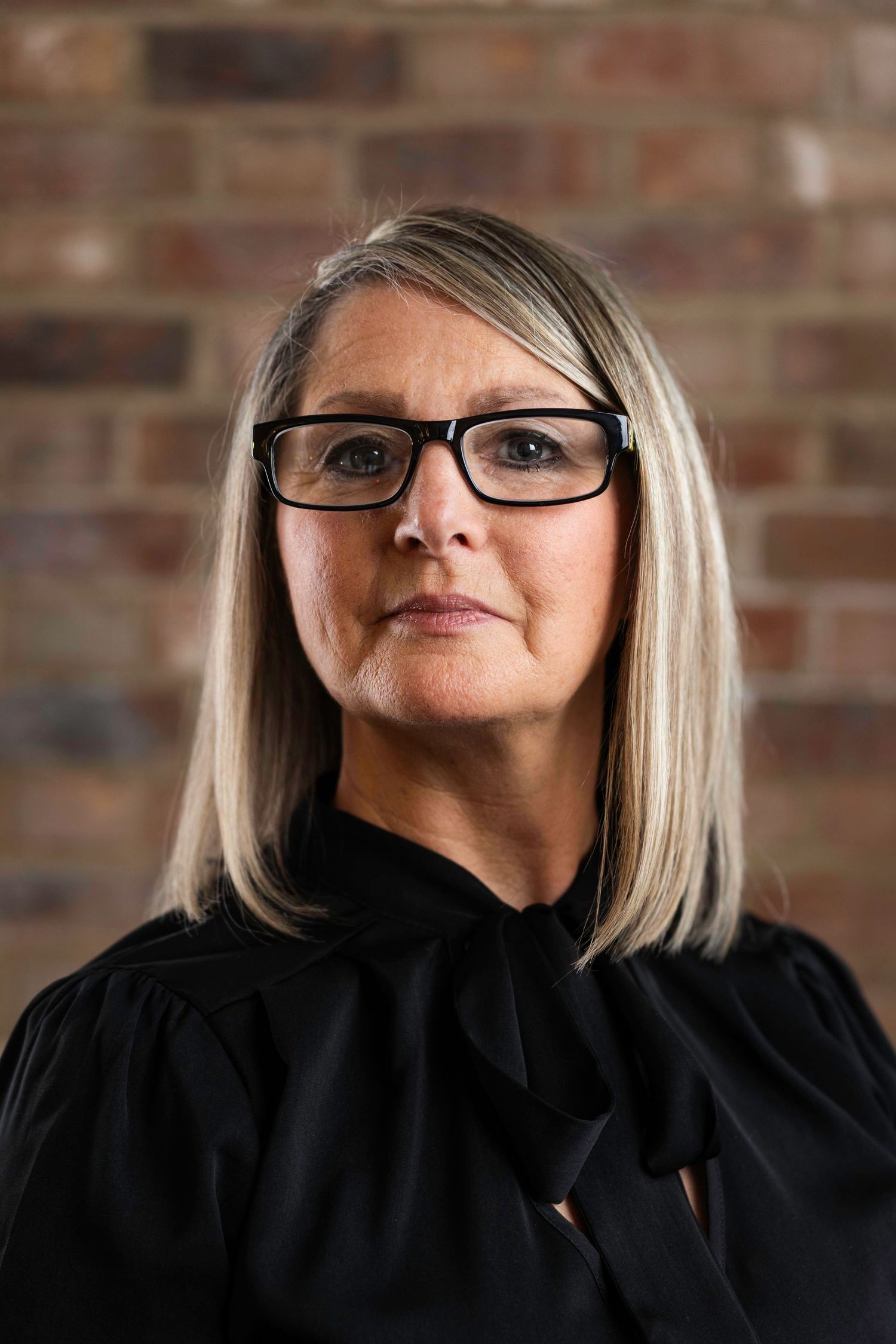 Professional portrait against brick wall showing blonde lady wearing black-rimmed glasses and dark high-necked blouse.