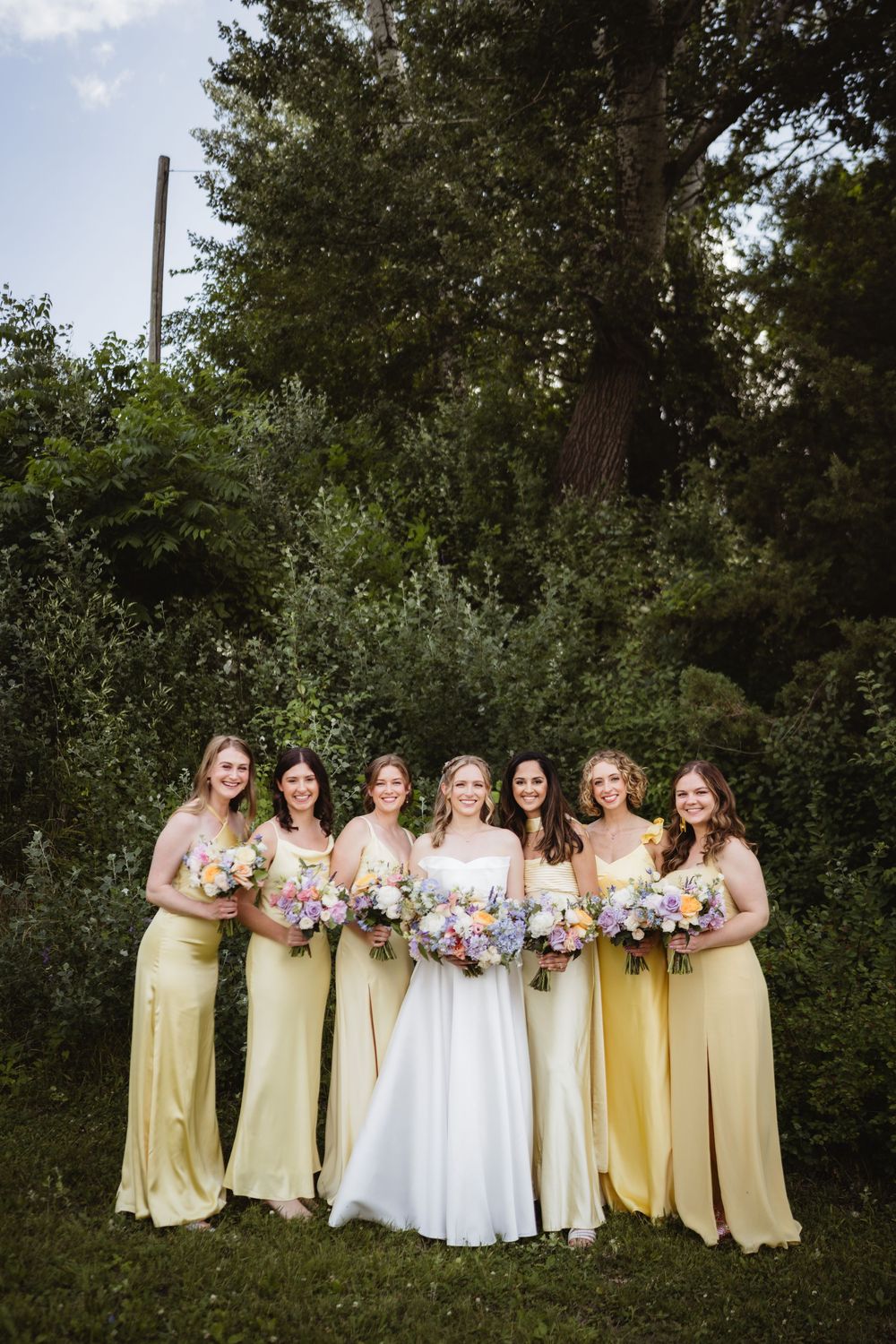 Yellow-gowns bridesmaids and bride pose for outdoor wedding photos in a garden setting with pink floral bouquets.