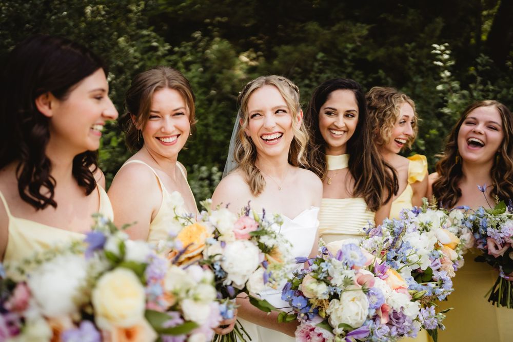 A group stands together outdoors in summer dresses holding colorful wildflower bouquets with purple and yellow blooms.