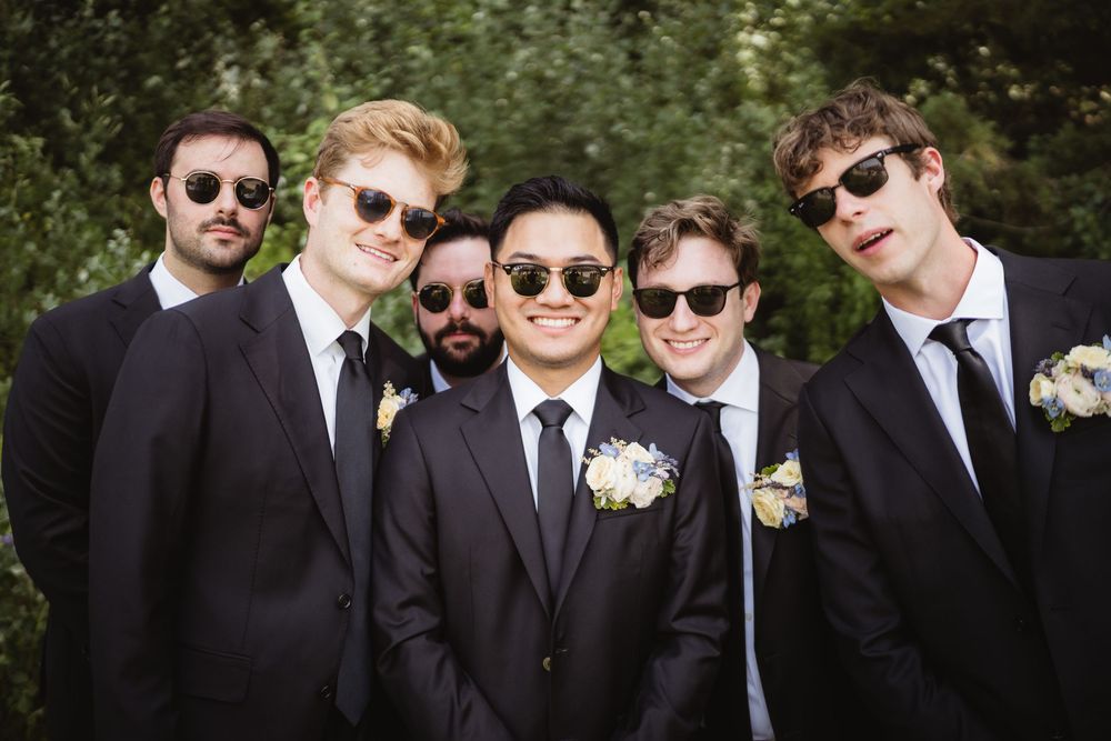 Five groomsmen in black suits and sunglasses pose together with boutonnieres at an outdoor wedding.
