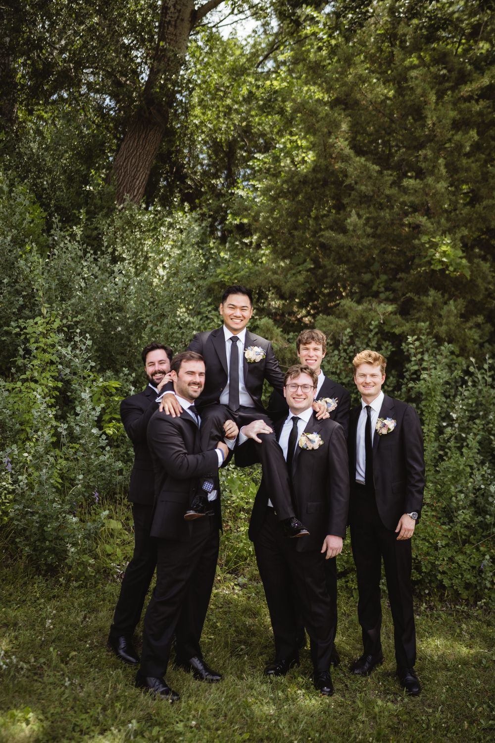 The groomsmen in black suits with boutonnieres stand in front of a lush green forest backdrop on a wedding day.