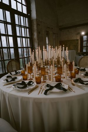Elegant table setting with white candles in glass bottles and black masks placed on white plates at each setting.