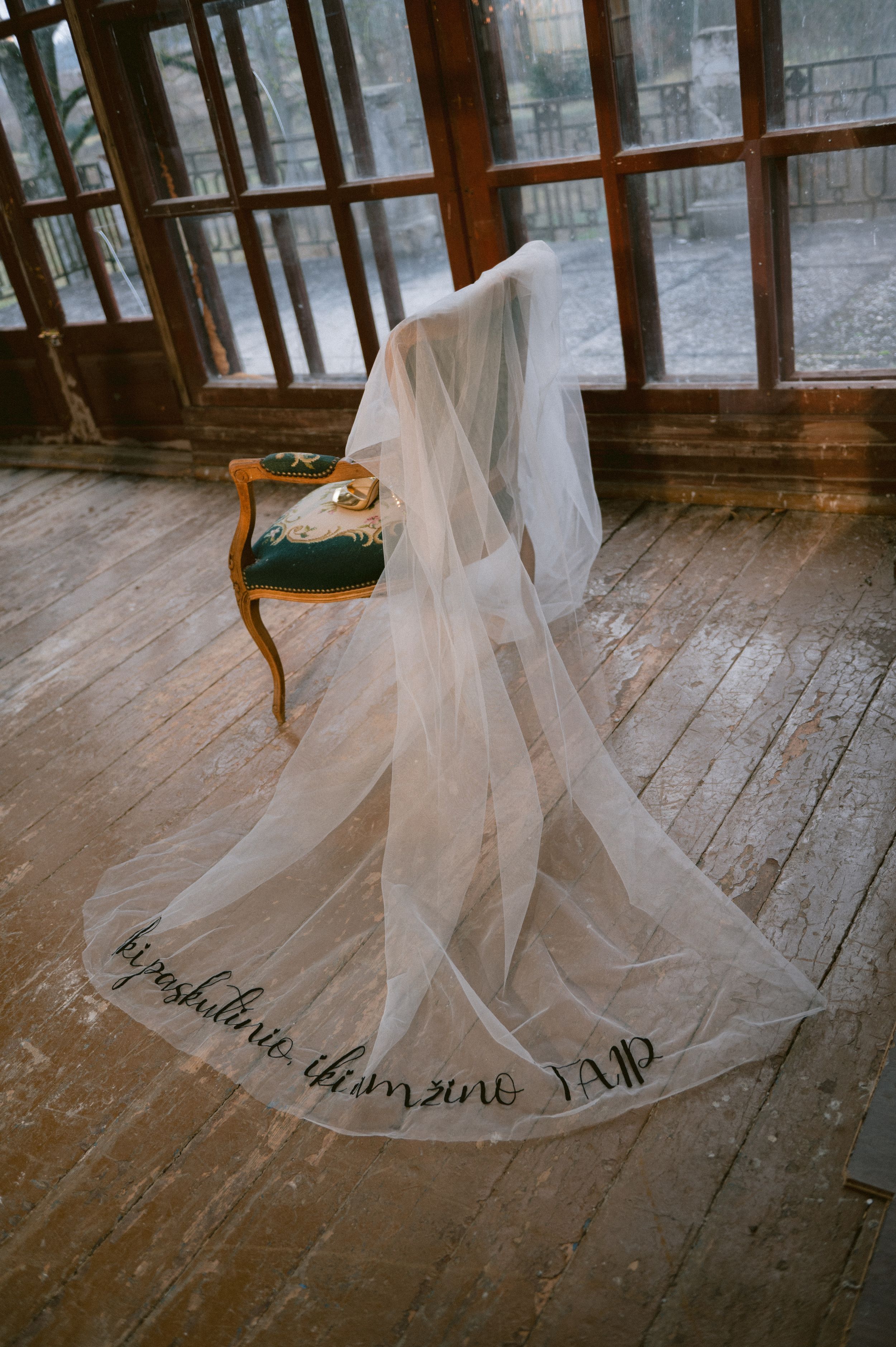 A flowing white wedding veil with text embroidery lying on a rustic wooden floor near vintage windows.