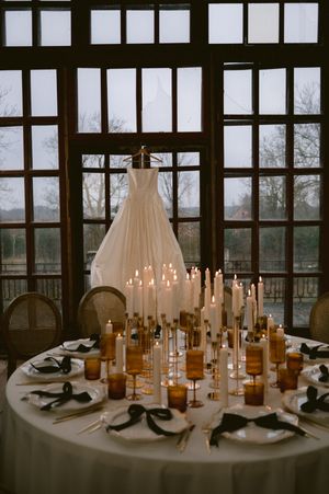 A white wedding dress hangs near a romantic table setting with numerous lit candles and place settings.