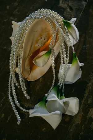 Pearl necklace and white calla lilies arranged on dark wooden surface with seashell.