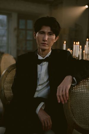 Moody portrait of person in formal black suit and white bow tie seated on vintage cane chair.