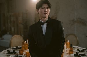A cinematic scene shows wine bottles and glassware on a table in a dimly lit room with dramatic lighting.