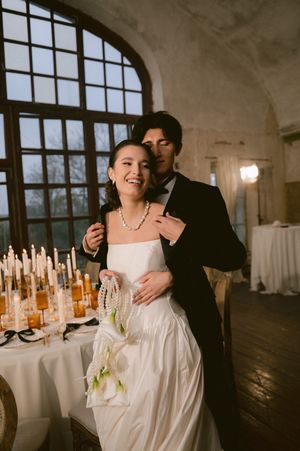 A romantic wedding couple embraces near an arched window with candles and elegant table settings in the background.