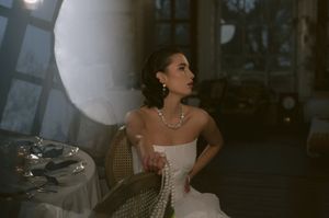 Woman in a white strapless gown and pearl necklace poses in a dimly lit room.