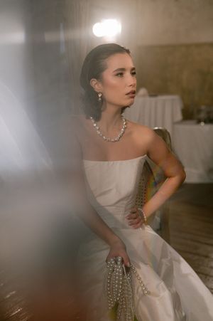 Elegant bride in strapless white gown and pearl jewelry poses in moody lighting against warm background.