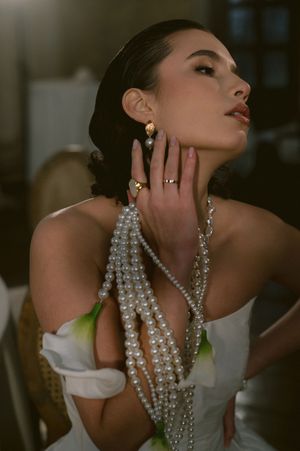 Elegant bride wearing dramatic pearl jewelry and off-shoulder dress in dramatic lighting.
