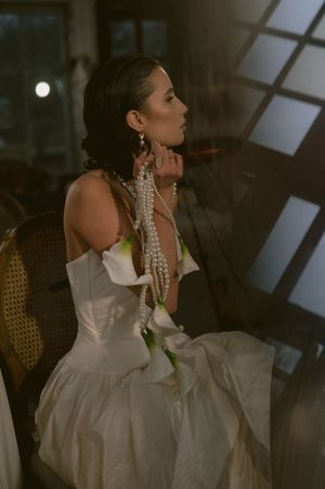 Woman in white gown and pearl necklace poses elegantly in dramatic lighting during a fashion photoshoot.