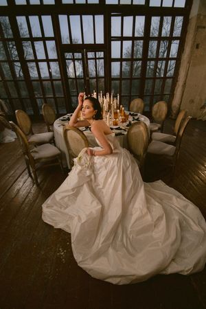 Bride in flowing white gown posed at candlelit table in rustic venue.