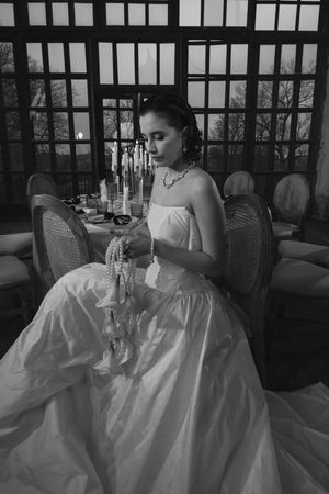 Black and white photo of bride in ballgown seated at candlelit table with window backdrop.