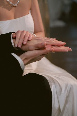 Close-up of delicate hands resting on white wedding gown fabric with pearl necklace visible.