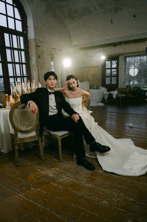 Wedding couple relaxes on vintage chairs in rustic industrial space with wooden floors and tall windows.