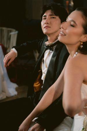 A bride and groom in formal wedding attire share a tender intimate moment in dramatic lighting against a dark background.