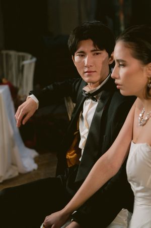 A formal couple in wedding attire sit together in a dimly lit venue, wearing a tuxedo and white gown with elegant details.