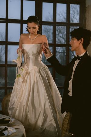 A bride in a voluminous white gown sits elegantly in a room with large windows during golden hour.