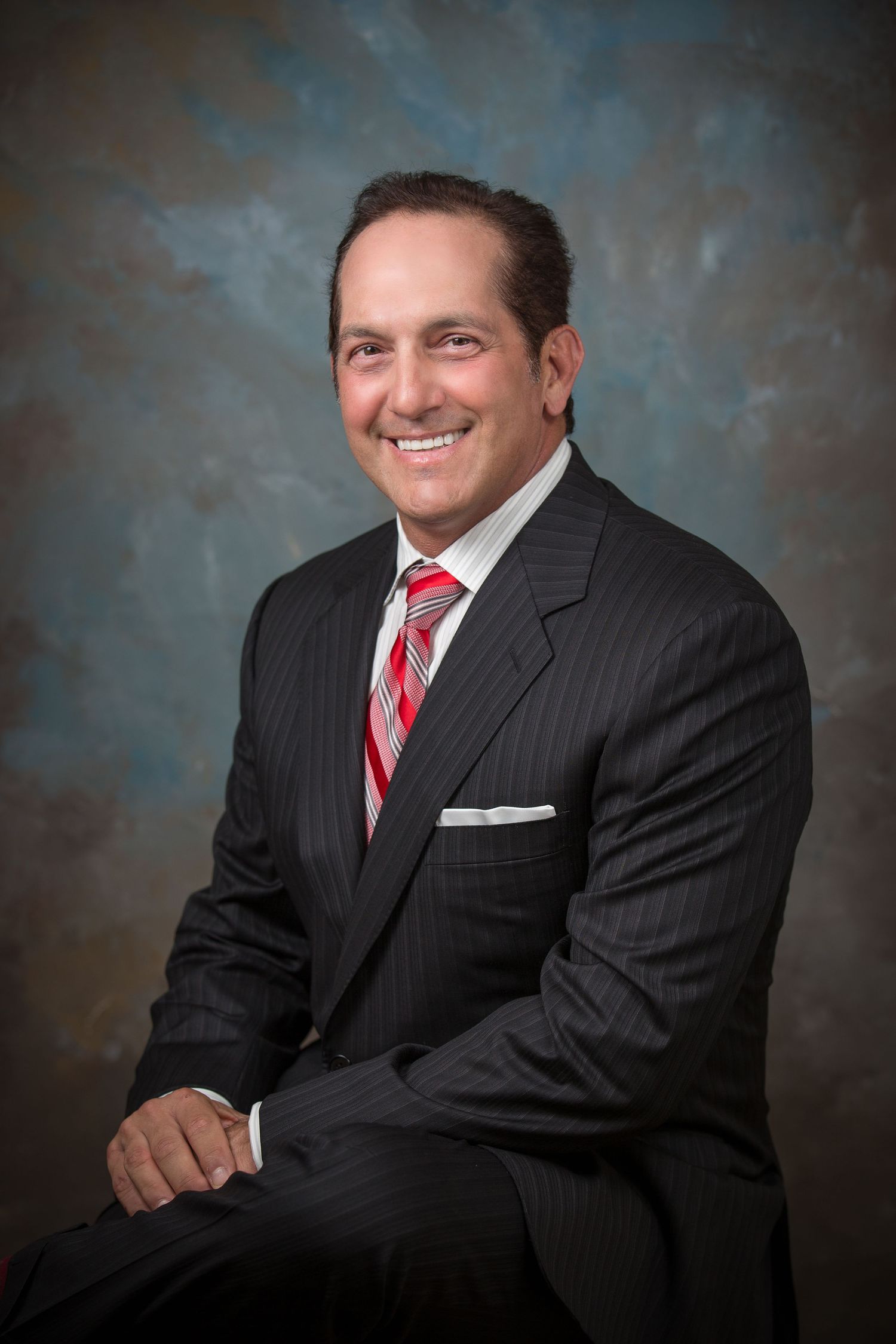 Professional headshot of a person in a pinstriped suit with red tie against a blue backdrop.