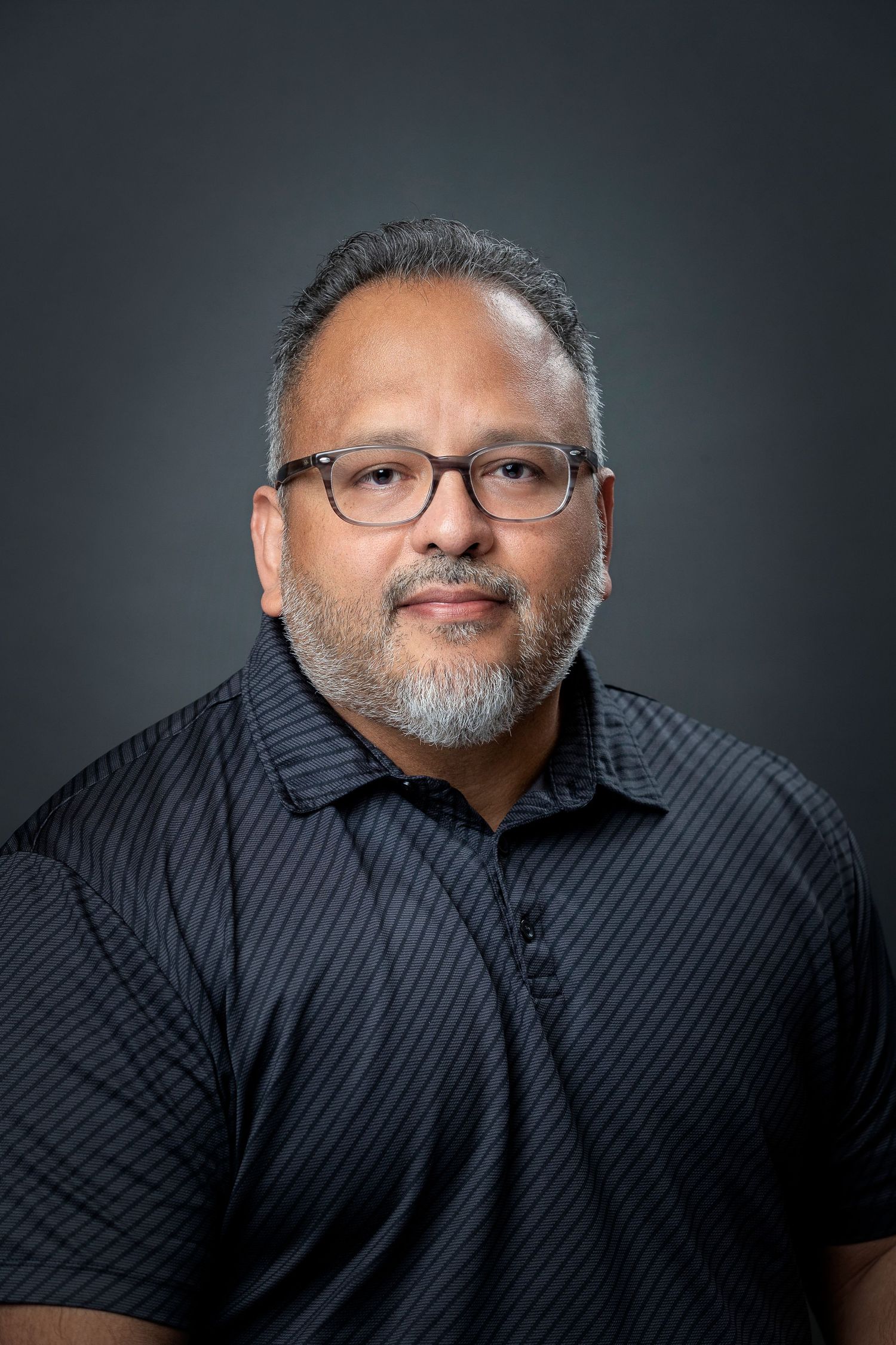 Professional headshot of a person in glasses and black polo against dark background.