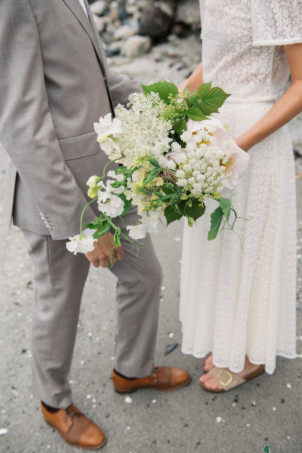Close-up of wedding attire details showing brown leather shoes, a gray suit, and a white dress with fresh floral bouquet.