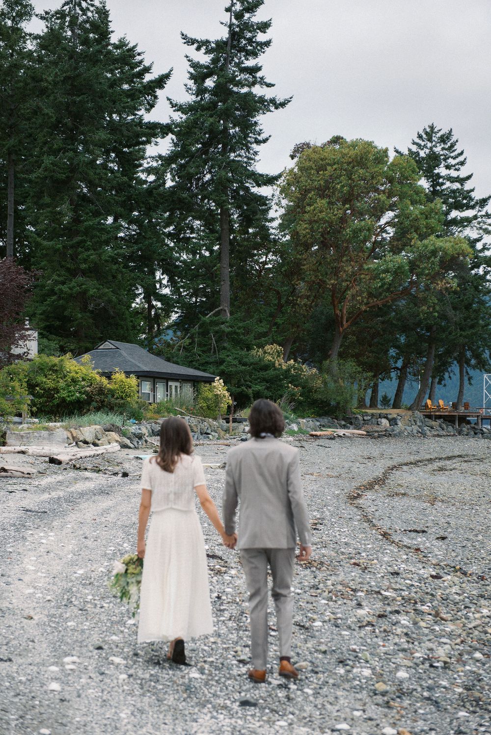 Couple walking on rocky beach path with evergreen trees in background on overcast day.