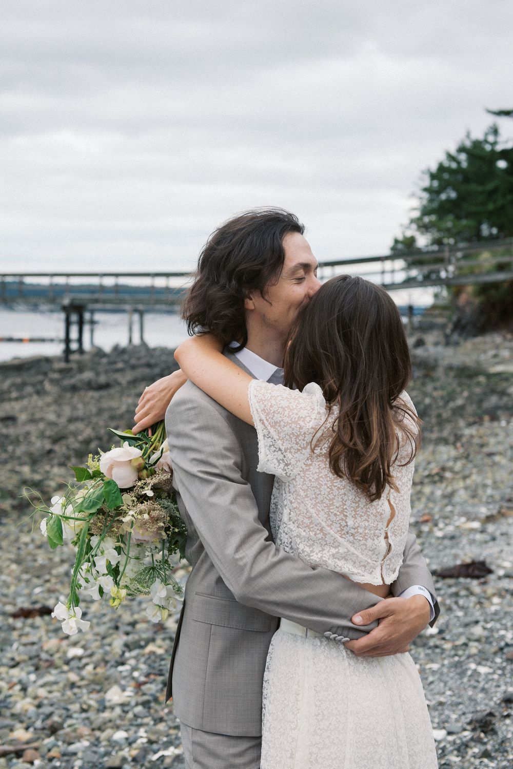 Intimate wedding moment captured on rocky beach with white and green bouquet details.