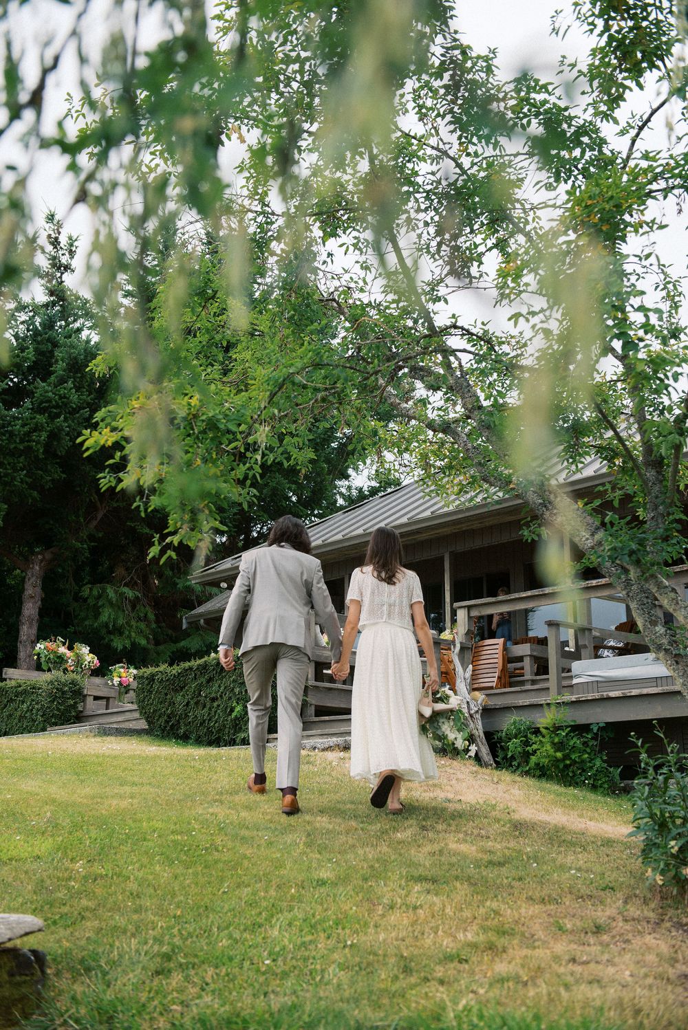Couple walking uphill on grassy slope with trees and rustic building in background.