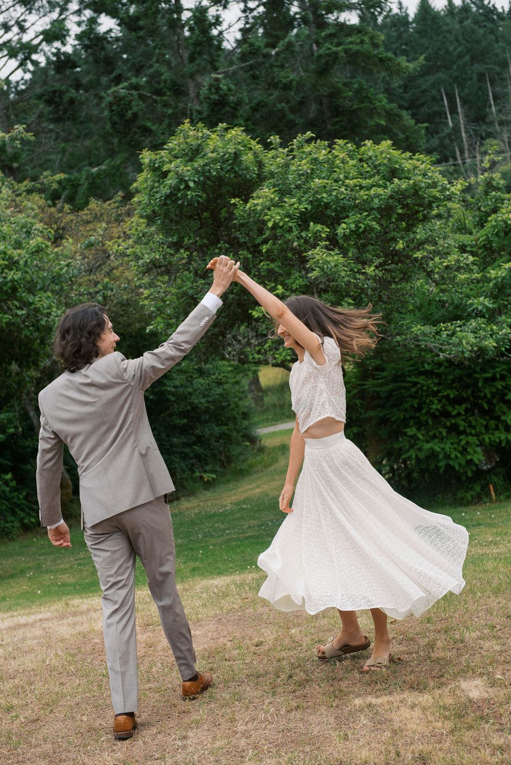 A couple dancing and twirling playfully in a grassy meadow with trees in the background.
