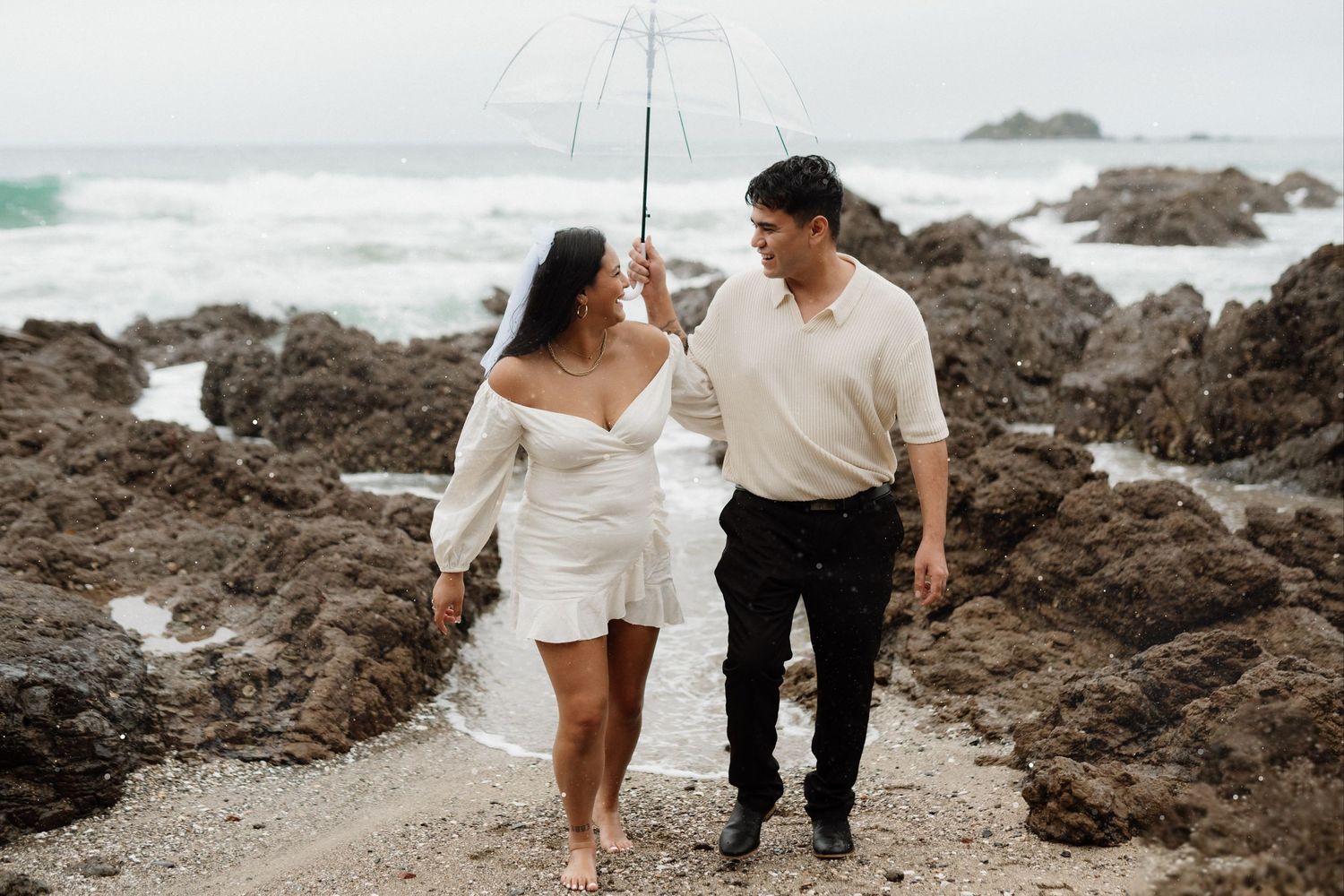 A couple in white and black attire walking along a rocky beach coastline at sunset.