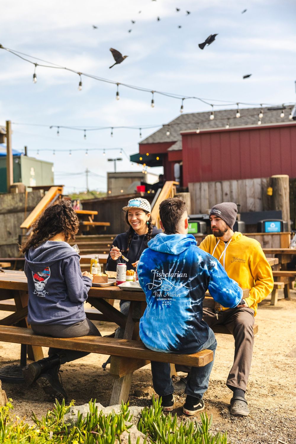 People gather at outdoor picnic tables in a rustic setting with string lights overhead and gravel ground below.