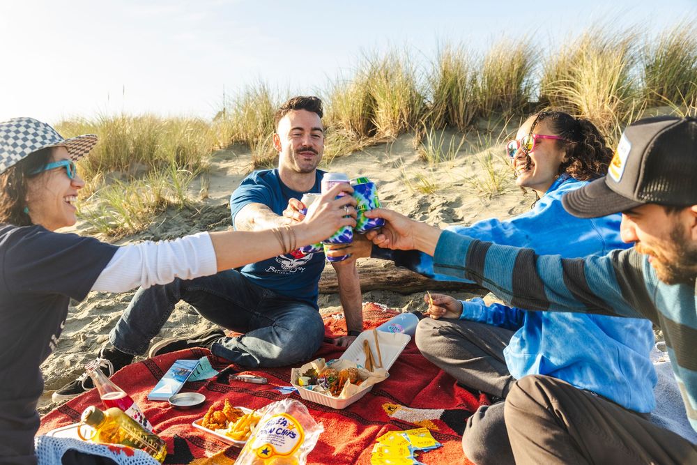 Group enjoying a picnic spread with blankets and snacks on a beach during golden hour.