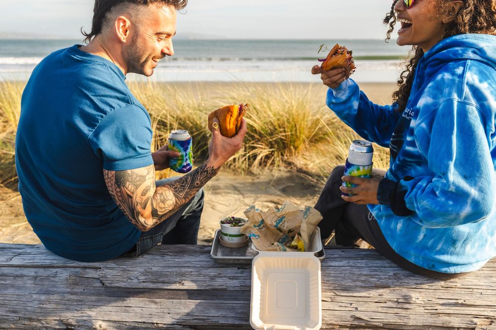 People enjoying drinks and snacks at a sunny beach setting with blankets and picnic supplies.