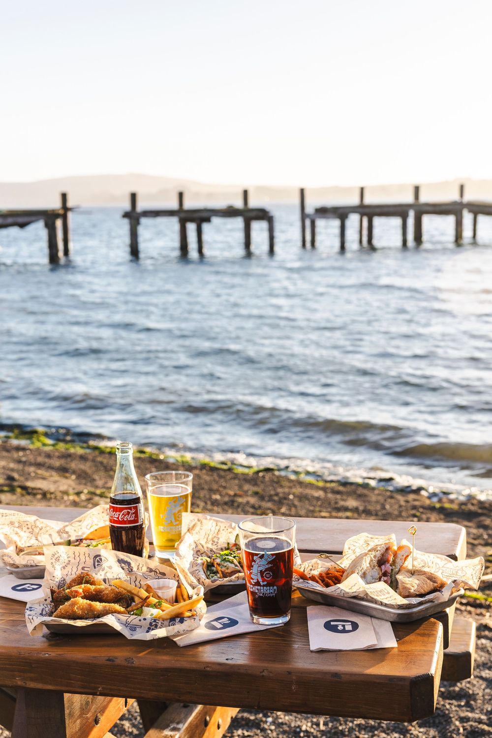 Beachside picnic spread with food and drinks on a wooden table overlooking the ocean and pier.