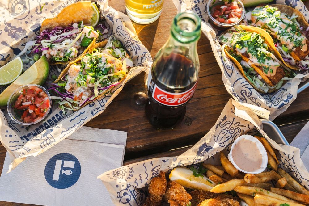 Casual dining spread with tacos, fries and a Coca-Cola bottle on a wooden table with foil-wrapped items.