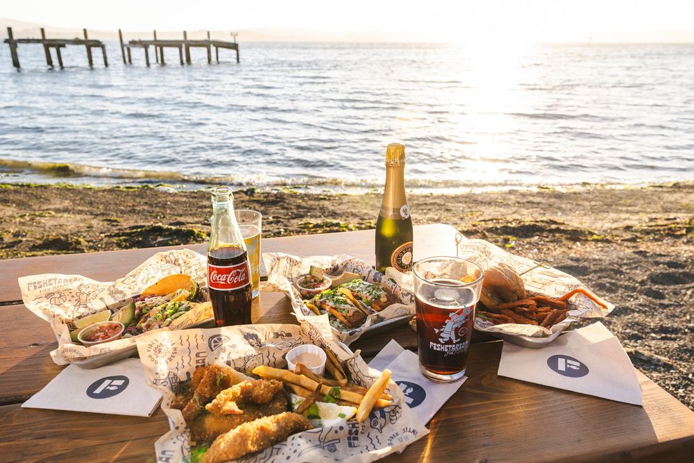 Beachside picnic setup with wine bottles, food platters and ocean view at sunset.