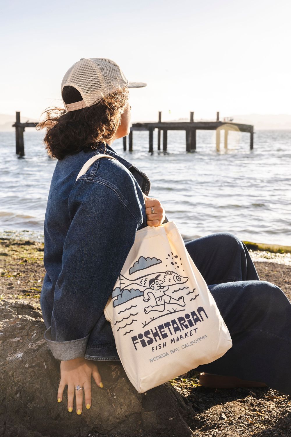 Person sitting by the ocean holding a tote bag with fishing-themed artwork at sunset.