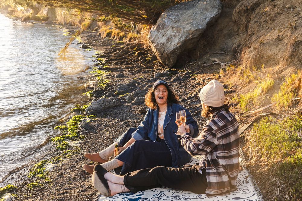 Two people enjoying wine and a picnic under a rocky cliff by the water at sunset.