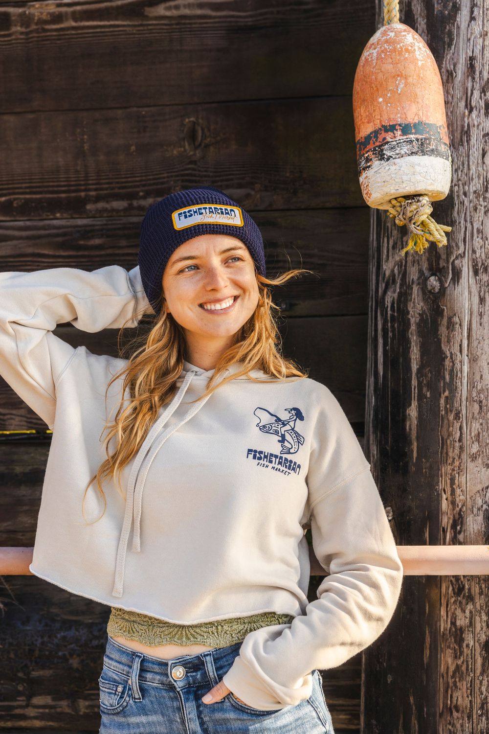 Person wearing a white sweatshirt and blue beanie posing against a rustic wooden wall.